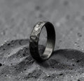 Hand-forged zirconium wedding band with black hammered finish, standing upright on dark surface