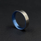 A metallic ring with a brushed silver exterior and a polished blue interior, standing upright on a dark textured surface.