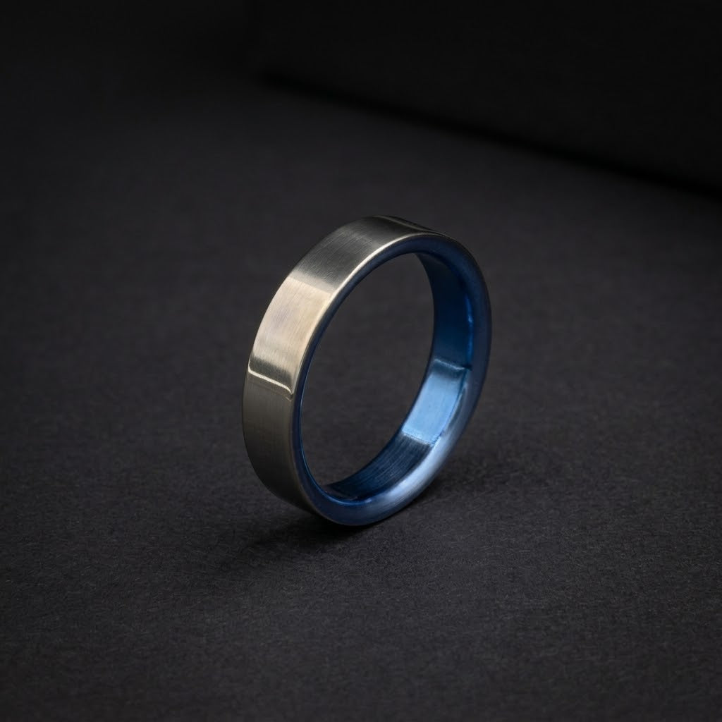 A minimalist metallic ring with a brushed silver exterior and a polished blue interior, standing upright on a dark textured surface.