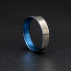 A brushed silver metal ring with a vibrant blue interior, standing upright on a dark textured surface.