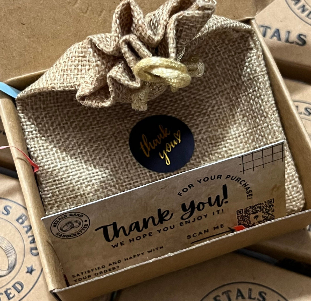 A small brown hessian bag with a gold "thank you" sticker. The bag is sitting in a brown cardboard box with a thank you card that says "Thank you! We hope you enjoy it! Scan me" and a QR code.