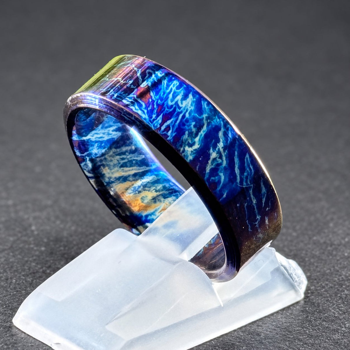 A blue and white titanium ring with a swirling pattern. The ring is on a white display stand.