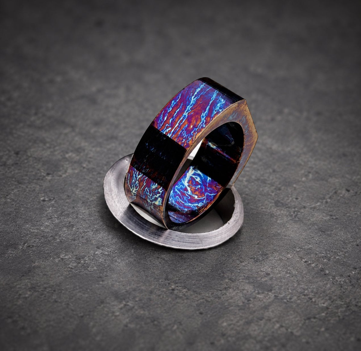 A hexagonal shaped ring with a blue, purple and red iridescent finish. The ring is sitting on a silver surface.