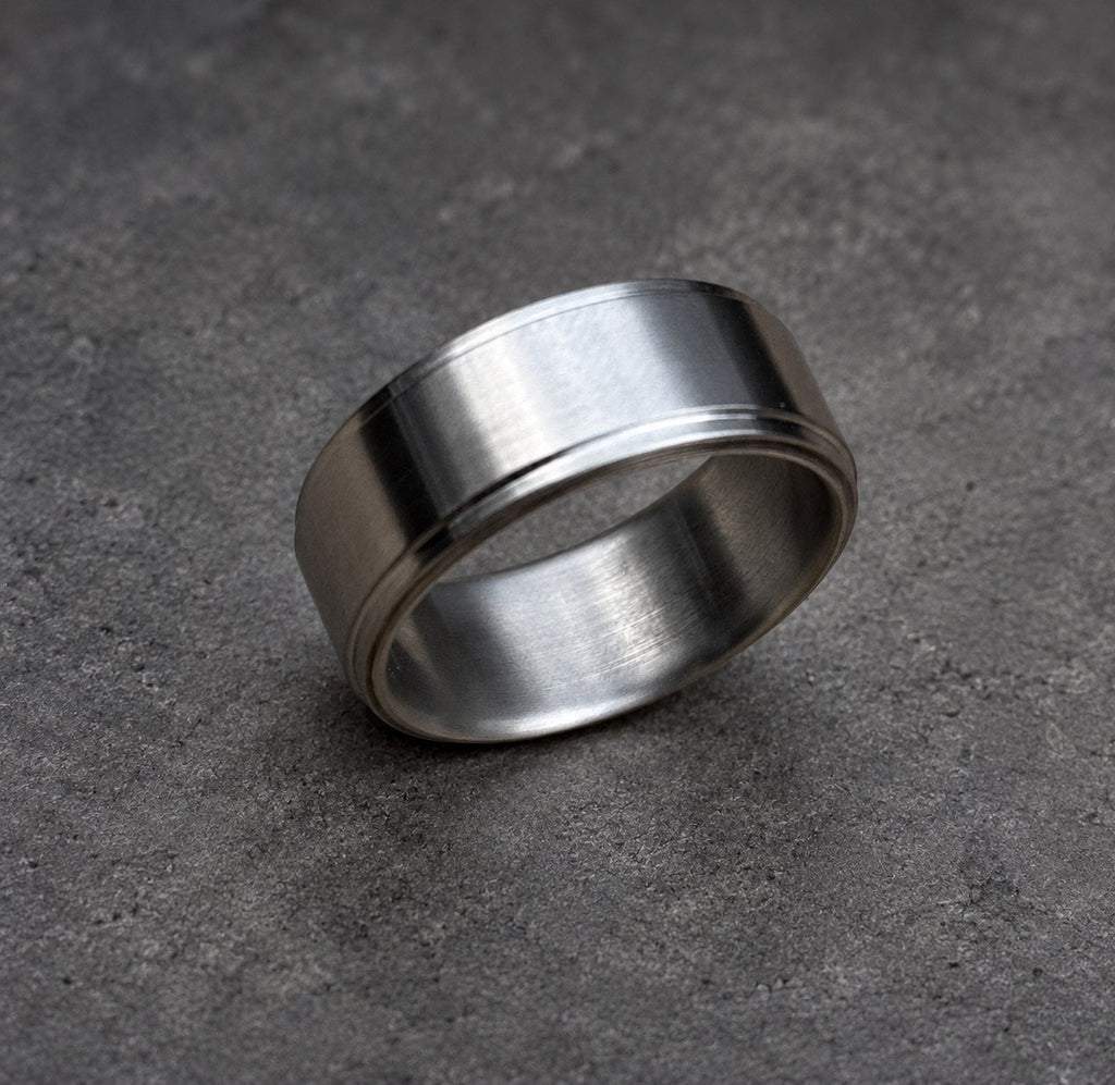 A brushed metal ring with a flat top and a grooved edge. The ring has a simple, modern design and a matte finish. The ring is a silver color and is set against a brown background.