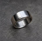 A brushed metal ring with a flat top and a grooved edge. The ring has a simple, modern design and a matte finish. The ring is a silver color and is set against a brown background.