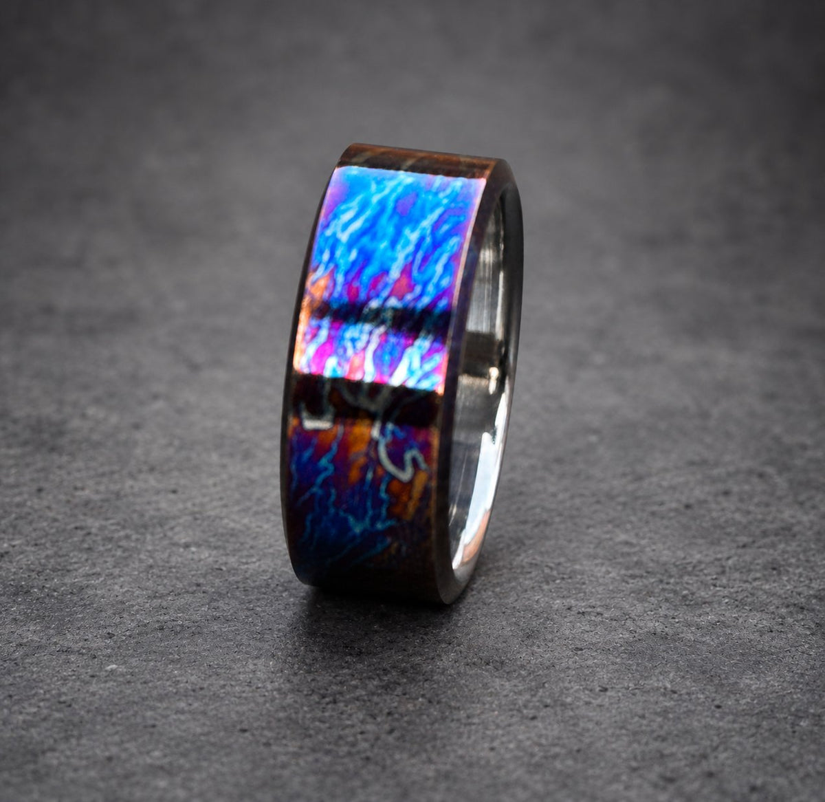 A wide, black band ring with a blue, purple and orange iridescent finish. The ring has a swirling pattern.