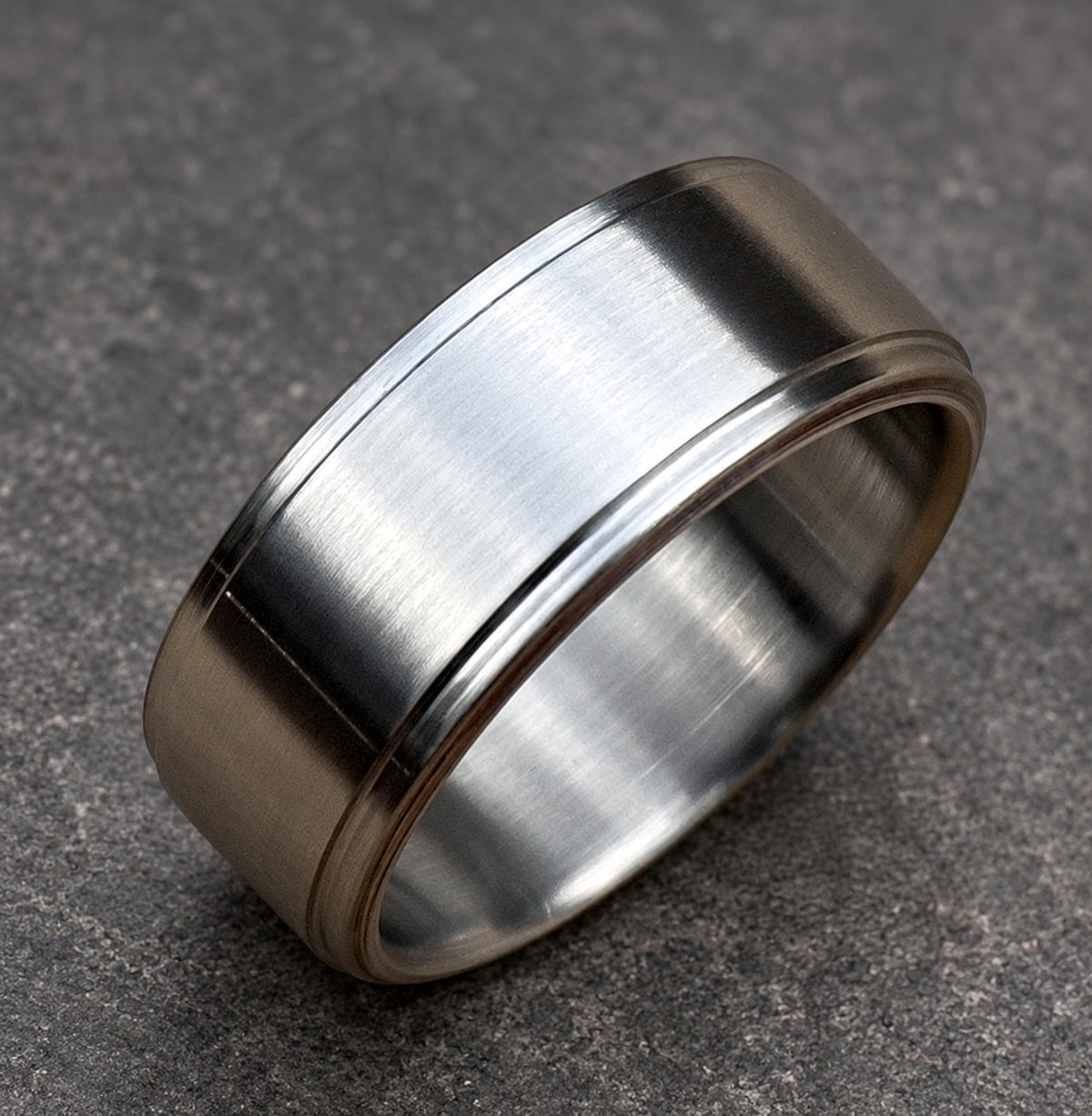 A brushed silver ring with a flat top and a grooved edge. The ring has a simple, modern design and a matte finish. The ring is likely made of stainless steel or a similar metal.