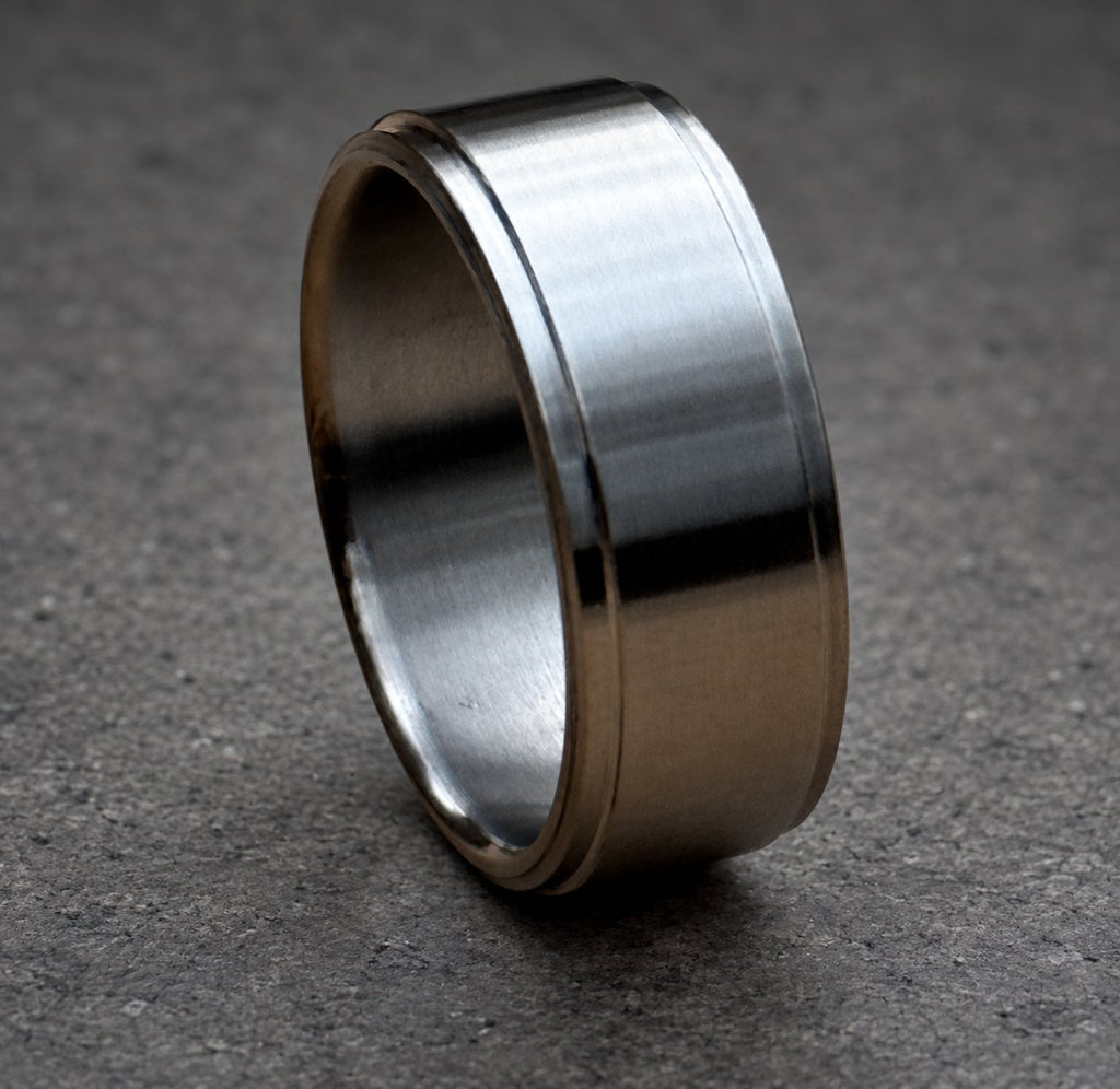 A brushed metal ring with a flat top and bottom, and a slightly rounded edge. The ring has a simple, minimalist design and a matte finish. The ring is shown on a neutral background.