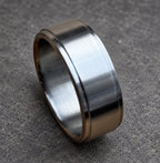 A close-up of a silver ring. The ring has a flat, wide band with a slightly raised edge on both sides. The ring is made of a shiny, metallic material.