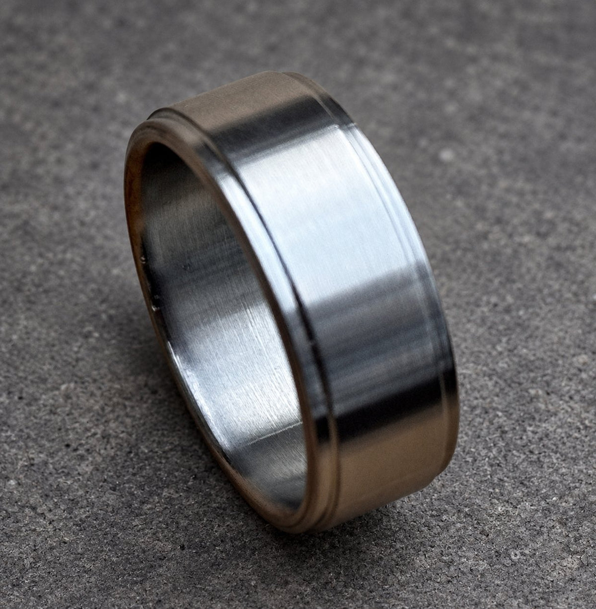 A close-up of a silver ring. The ring has a flat, wide band with a slightly raised edge on both sides. The ring is made of a shiny, metallic material.