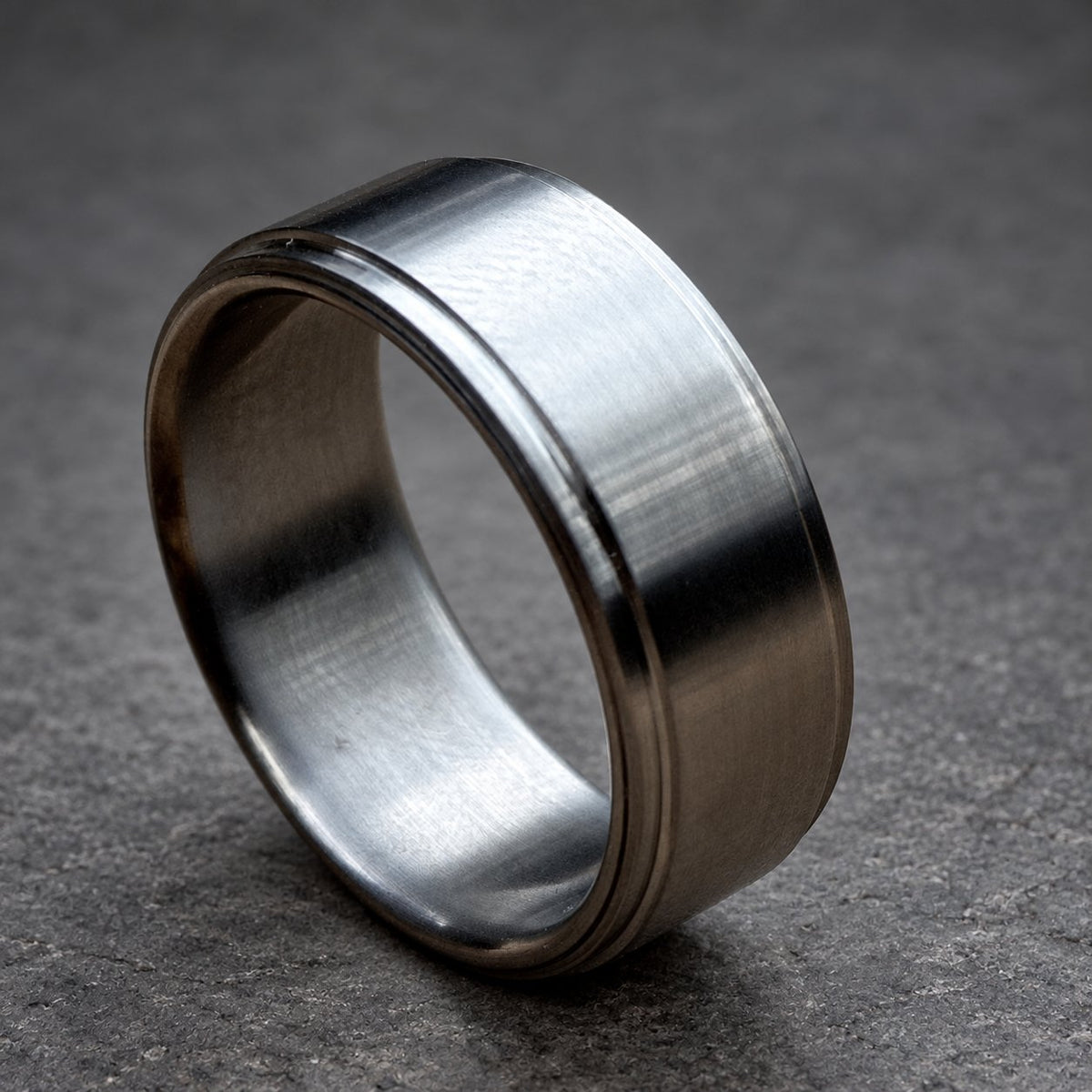 A brushed silver ring with a flat top and a grooved edge. The ring has a simple, modern design and a matte finish. The ring is likely made of stainless steel or a similar metal, view 2