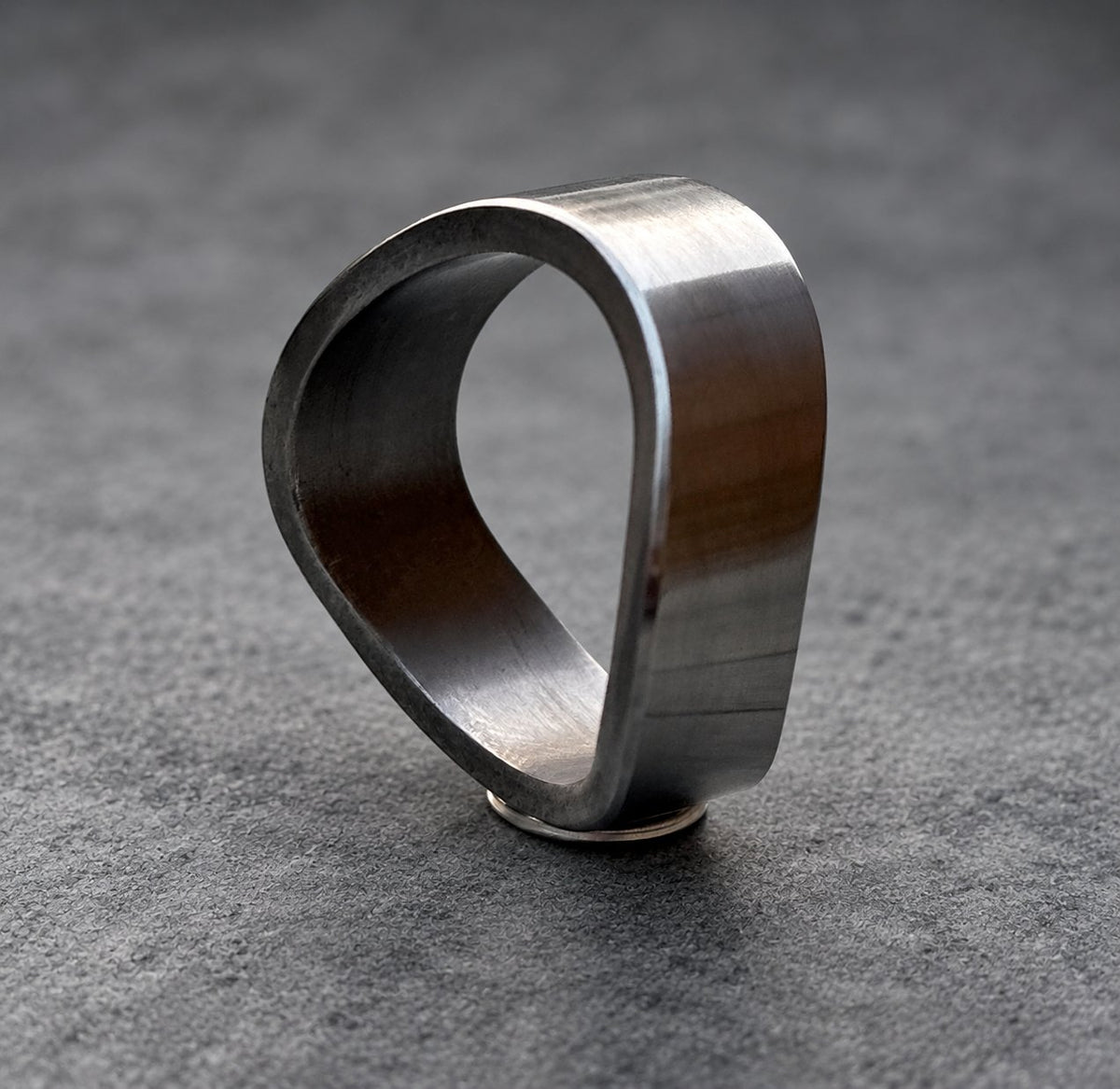 A dark gray, metal ring with a unique, asymmetrical design. The ring is made of a single piece of metal that is bent into a shape resembling a teardrop.