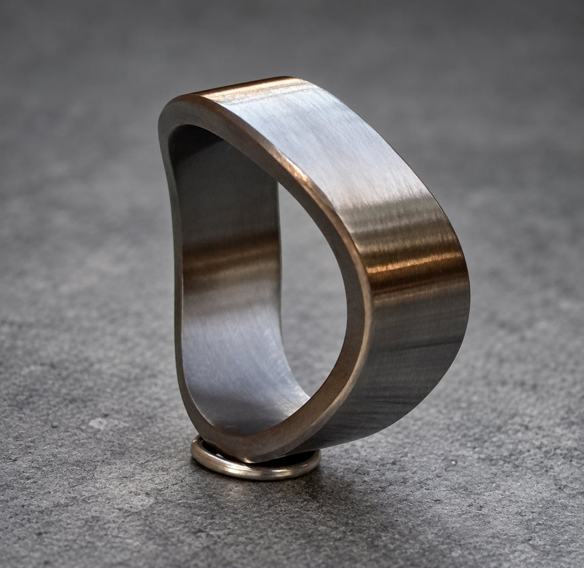 A dark gray metal ring with a unique, asymmetrical design. The ring is smooth and has a brushed finish.