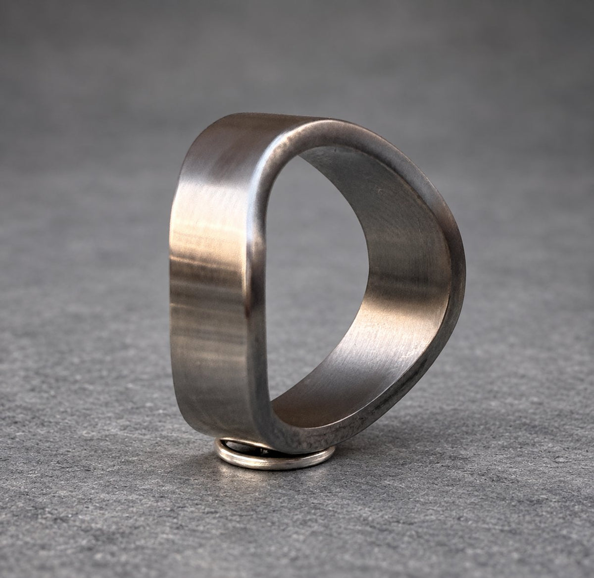 A silver ring with a unique, asymmetrical, rounded, and slightly curved design. The ring is made of metal and has a brushed finish.
