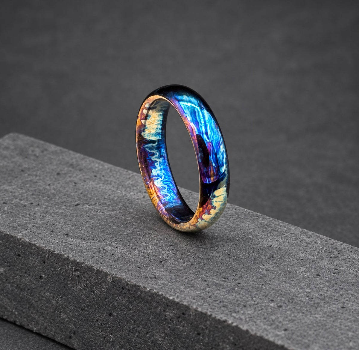 A close-up of a colorful, iridescent metal ring. The ring has a blue, purple, and orange color scheme. The ring is sitting on a wooden surface.