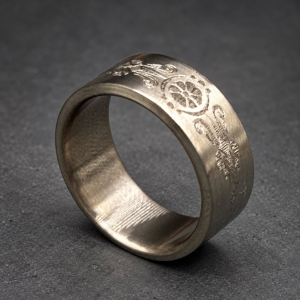 A silver ring with an engraved floral and geometric pattern. The ring has a wide band and a matte finish. The design is repeated around the band. The ring is displayed on a gray background.