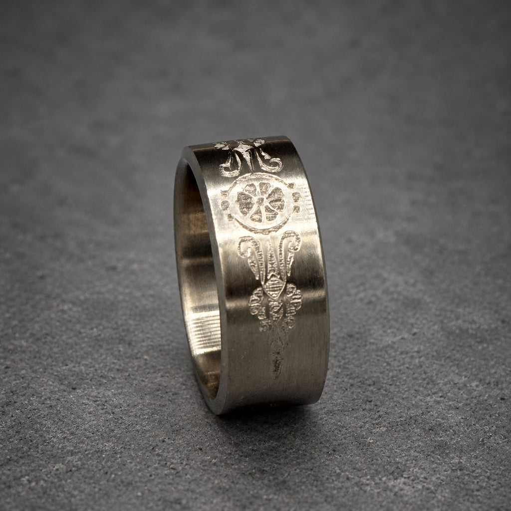 A silver ring with an engraved design. The ring features a central floral or wheel-like motif, flanked by ornate, symmetrical patterns. The ring's surface has a matte finish, and the design is etched into the metal.