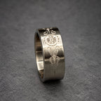A silver ring with an engraved design. The ring features a central floral or wheel-like motif, flanked by ornate, symmetrical patterns. The ring's surface has a matte finish, and the design is etched into the metal.