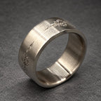 A silver ring with etched floral patterns. The ring features a wide band with a textured finish. The ring is made of metal and is shown on a gray surface.