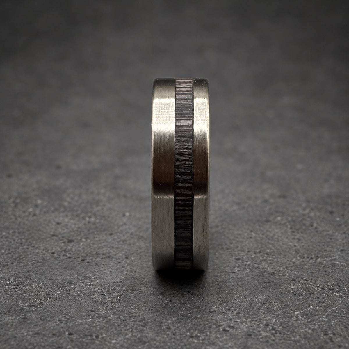 A silver ring with a dark wood inlay is shown in close-up. The ring features a silver-colored metal band with a central inlay of dark, textured wood. The ring is set against a blurred wooden surface.