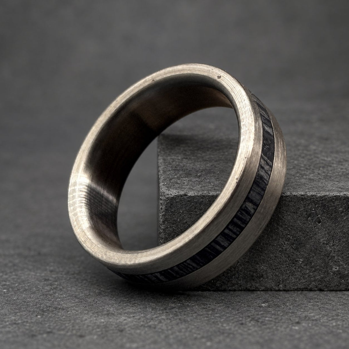A silver ring with a dark wood inlay. The ring has a brushed metal finish and a smooth, rounded shape. The wood inlay is a dark gray color and runs around the circumference of the ring.