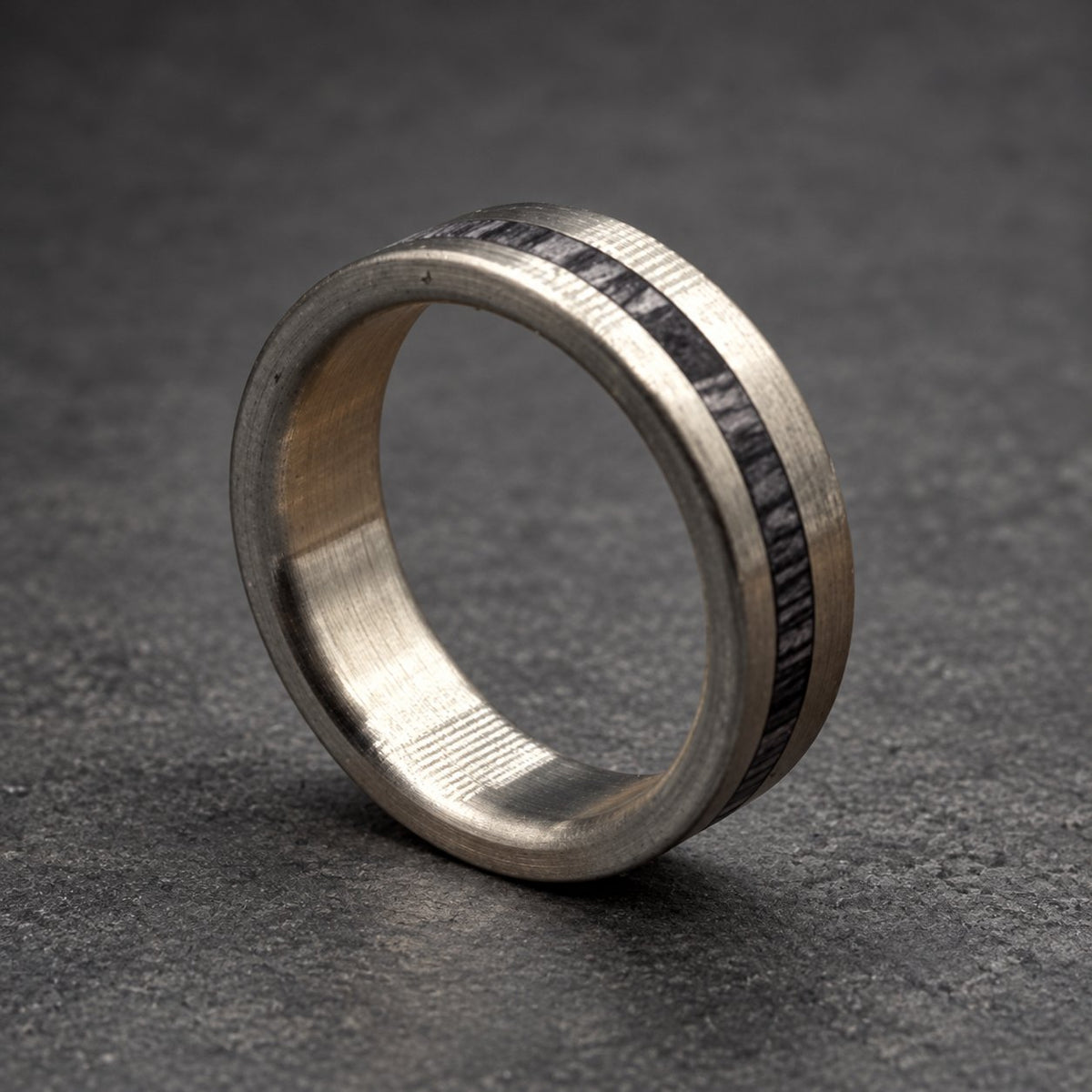 A silver ring with a dark, textured inlay. The ring is displayed on a wooden surface, highlighting its metallic finish and the contrast between the smooth metal and the textured inlay. The ring's design is simple and elegant.