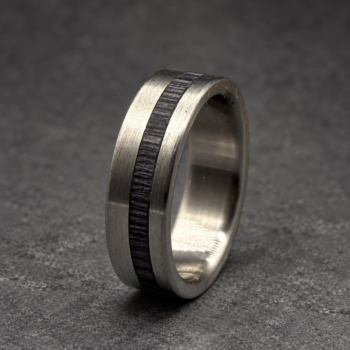 A silver ring with a dark wood inlay. The ring has a brushed metal finish and a flat profile. The dark wood inlay runs around the center of the ring, creating a striking contrast. The ring is a piece of jewelry.