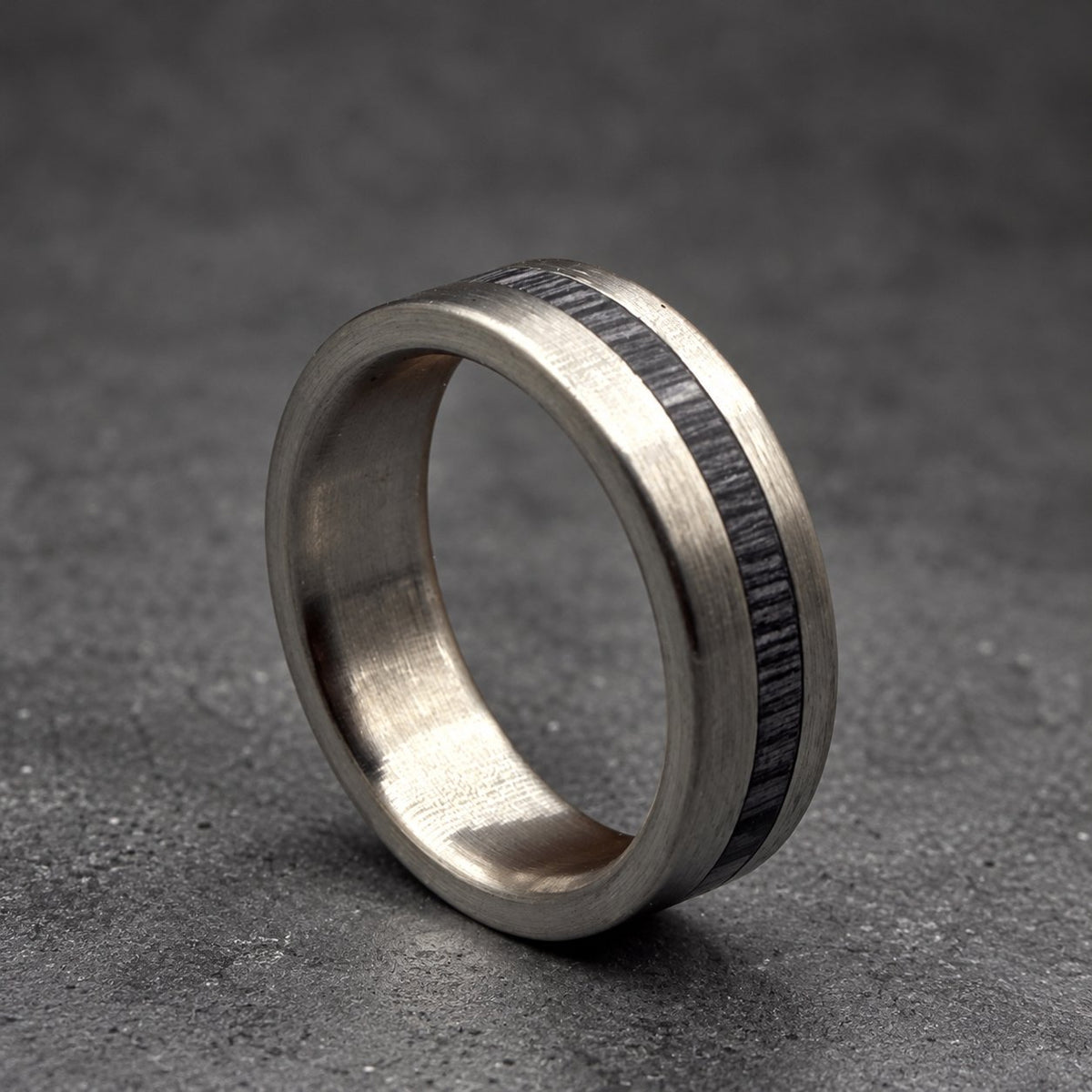 A silver ring with a dark wood inlay. The ring has a brushed metal finish and a smooth, rounded design. The wood inlay is a dark gray color and runs around the center of the ring.