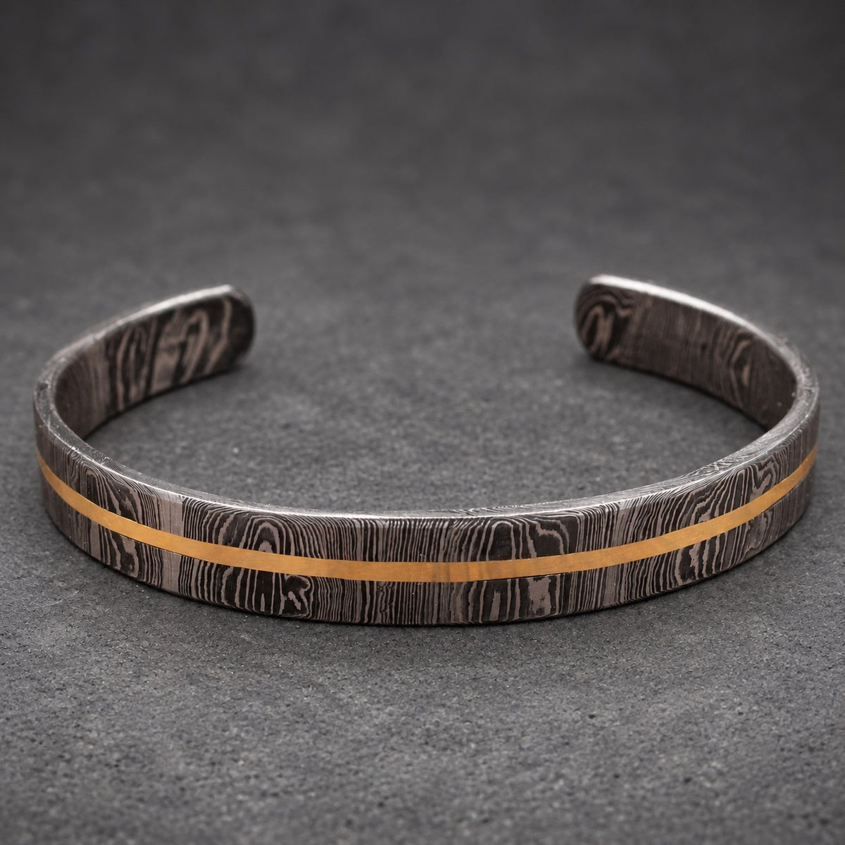 A Damascus steel cuff bracelet with a gold-colored stripe. The bracelet has a dark, patterned finish and a simple, open design. The gold stripe runs horizontally across the center of the bracelet.