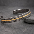 A Damascus steel cuff bracelet with a gold-colored stripe. The bracelet has a unique wood grain pattern and a polished finish. This open-ended bracelet is a stylish accessory.