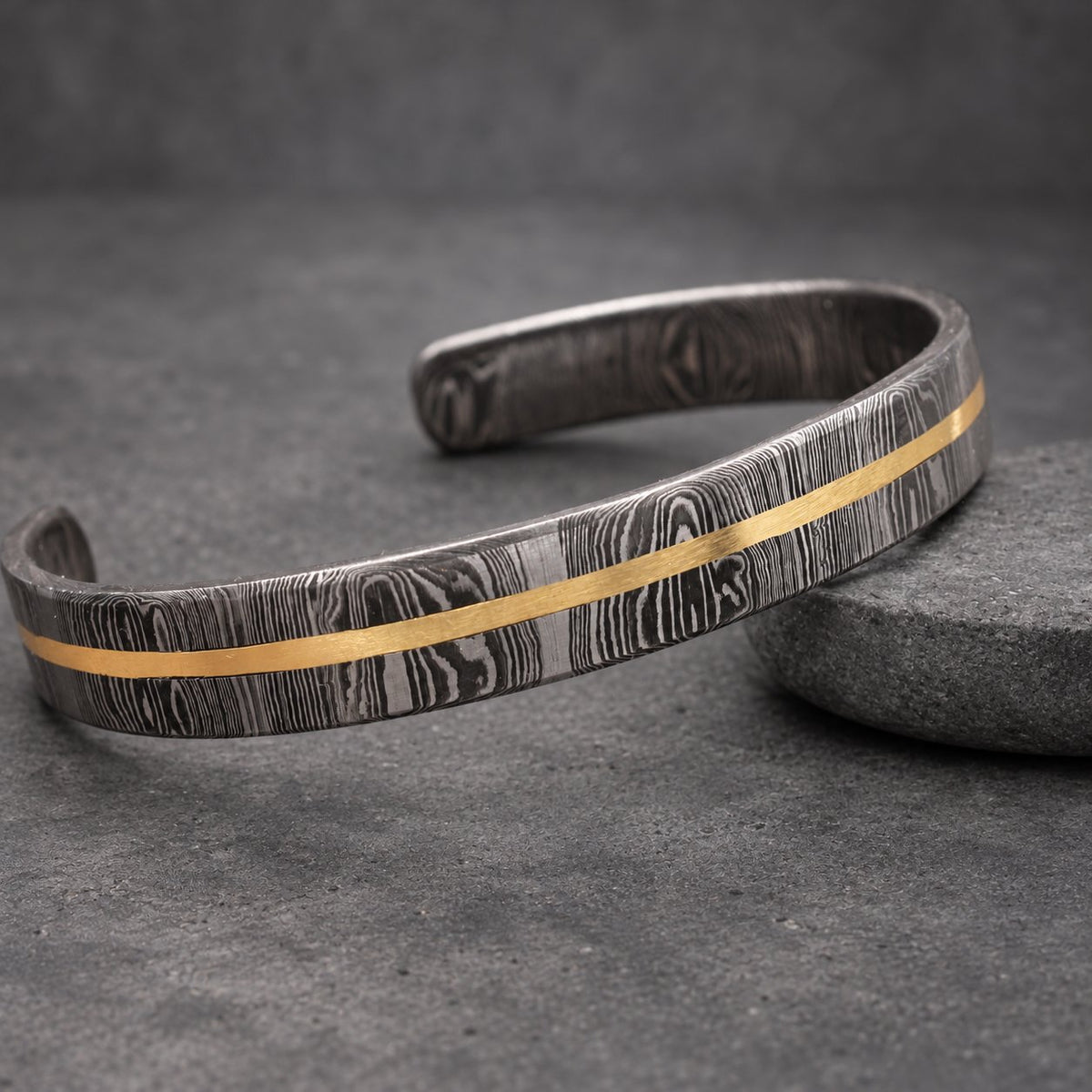 A Damascus steel cuff bracelet with a gold-colored stripe. The bracelet has a unique wood grain pattern and a polished finish. This open-ended bracelet is a stylish accessory.