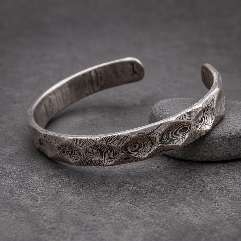 A dark gray, textured cuff bracelet with an open design. The bracelet has a distinctive pattern of swirling lines and geometric facets. It is displayed on a small wooden block. The bracelet's design allows for easy wear.