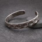 A dark gray, textured cuff bracelet with an open design. The bracelet has a distinctive pattern of swirling lines and geometric facets. It is displayed on a small wooden block. The bracelet's design allows for easy wear.