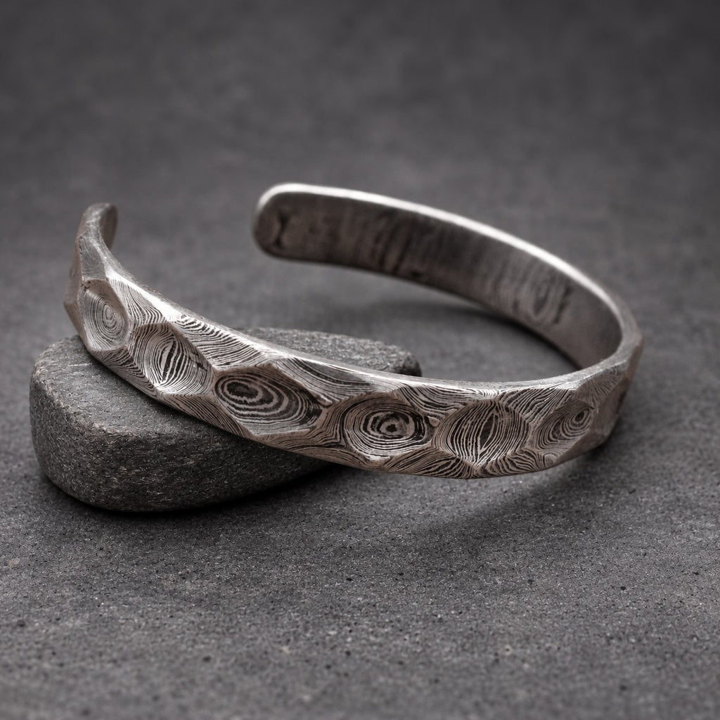 A silver cuff bracelet with a distinctive, textured pattern. The open-ended bracelet is made of metal and rests on a small wooden block. The surface of the bracelet has a swirling, organic design.