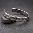 A silver cuff bracelet with a distinctive, textured pattern. The open-ended bracelet is made of metal and rests on a small wooden block. The surface of the bracelet has a swirling, organic design.