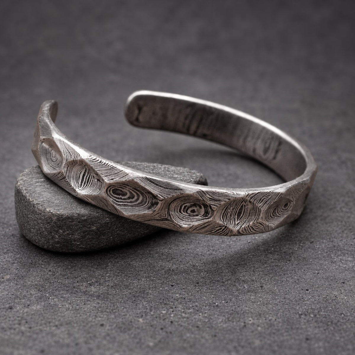 A silver cuff bracelet with a distinctive, textured pattern. The open-ended bracelet is made of metal and rests on a small wooden block. The surface of the bracelet has a swirling, organic design.