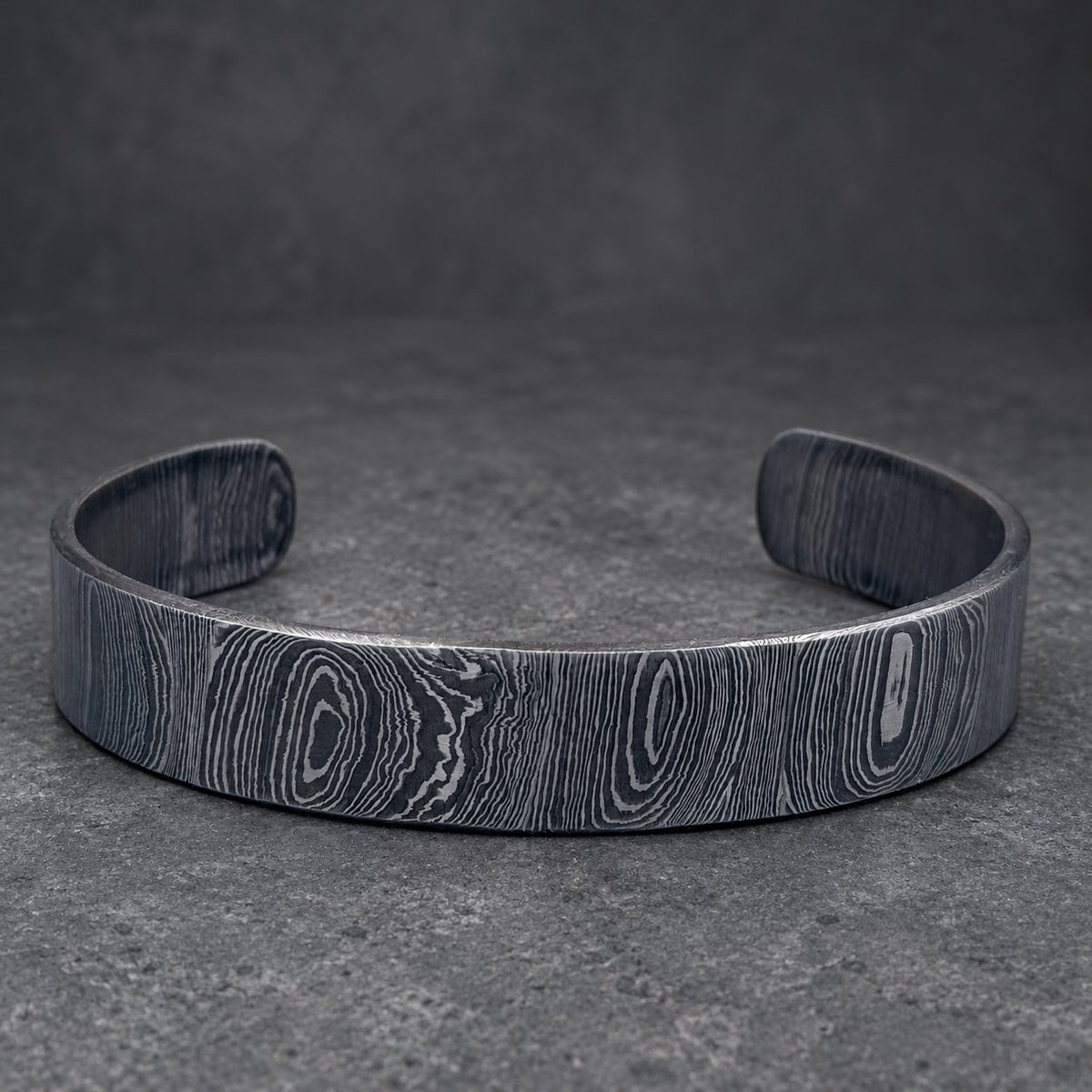 A dark gray metal cuff bracelet with a wood grain pattern. The open-ended bracelet is displayed on a light gray surface with a gold-colored decorative object in the background.