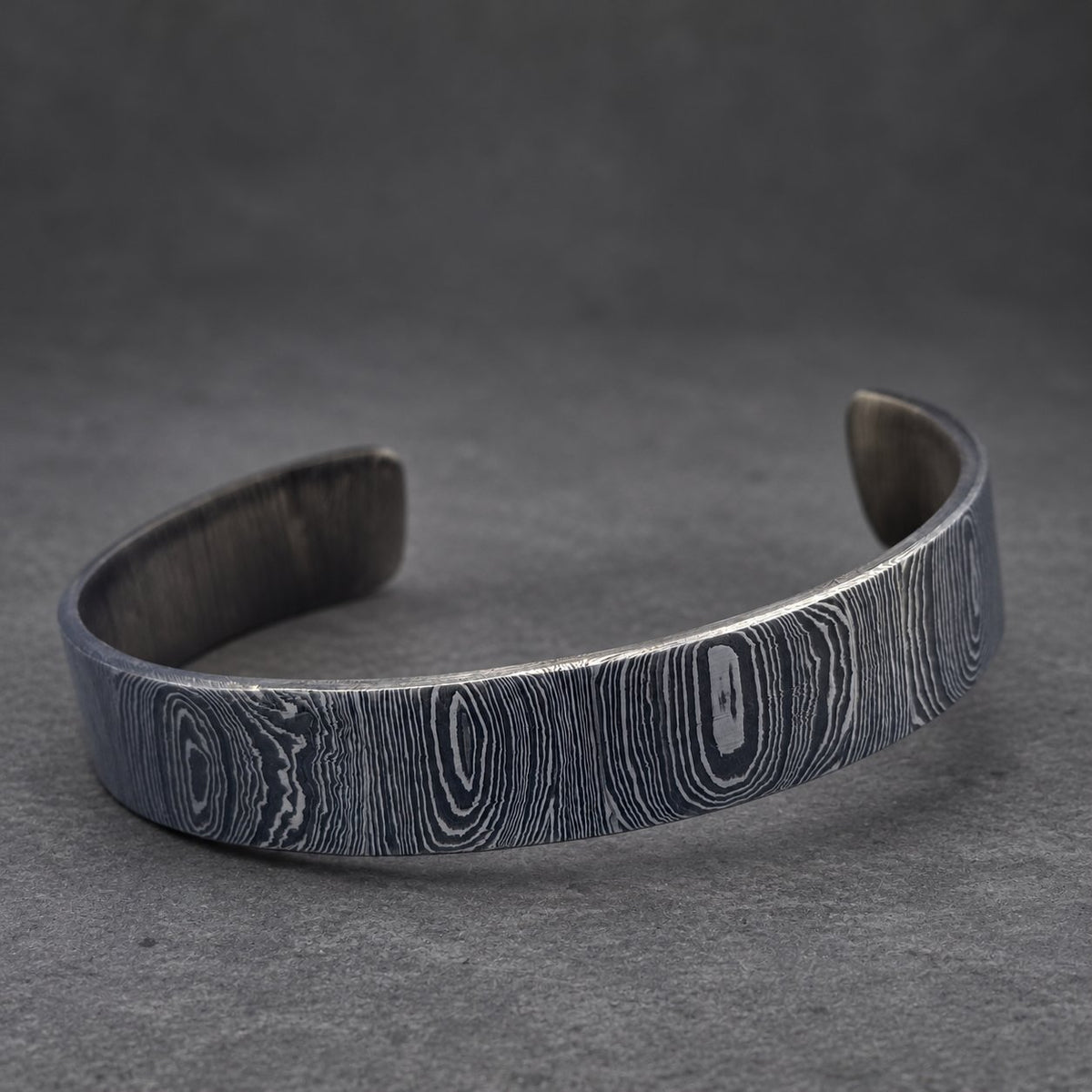 A dark gray metal cuff bracelet with a wood grain pattern. The bracelet is curved and open at the bottom. The background features a gold-colored, textured object.