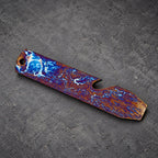 A multi-colored bottle opener with a unique pattern of blue, purple, and gold hues. The metal opener has a hole at one end and a bottle cap remover at the other. The design resembles a stormy sky.