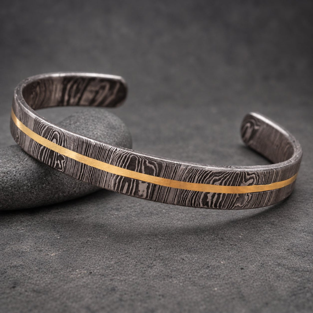 A Damascus steel cuff bracelet with a gold-colored stripe. The bracelet has a dark, patterned finish and a simple, open design. The gold stripe runs along the center of the bracelet.