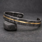 A Damascus steel cuff bracelet with a gold-colored stripe. The bracelet has a dark, patterned finish and a simple, open design. The gold stripe runs along the center of the bracelet.