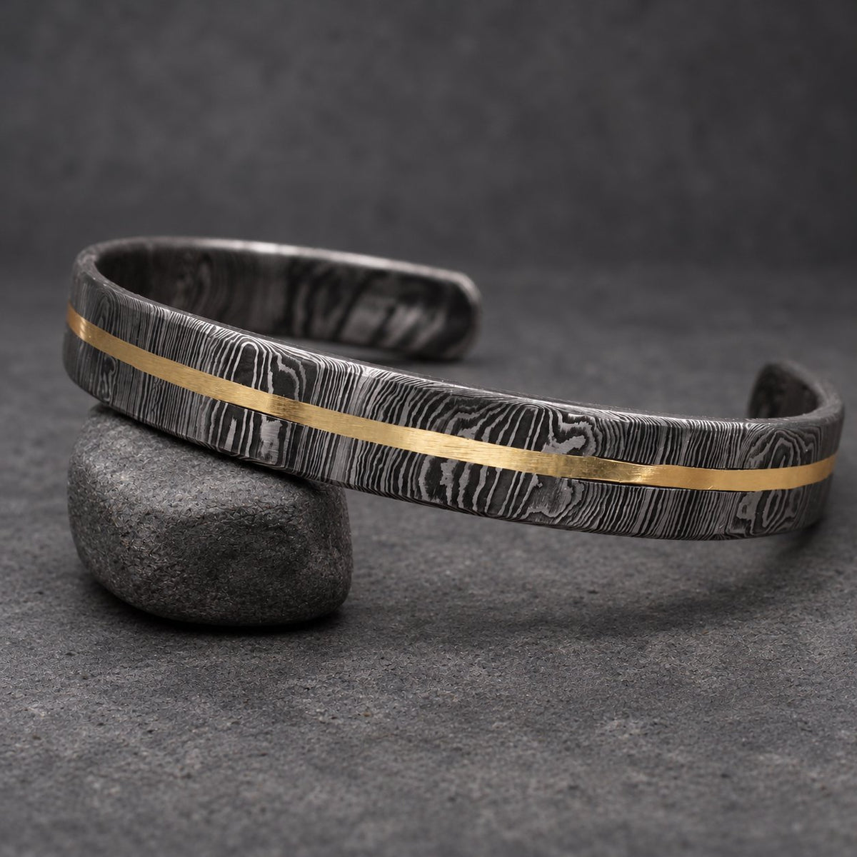 A Damascus steel cuff bracelet with a gold-colored stripe. The bracelet has a dark, patterned finish and a simple, open design. The gold stripe runs along the center of the bracelet.