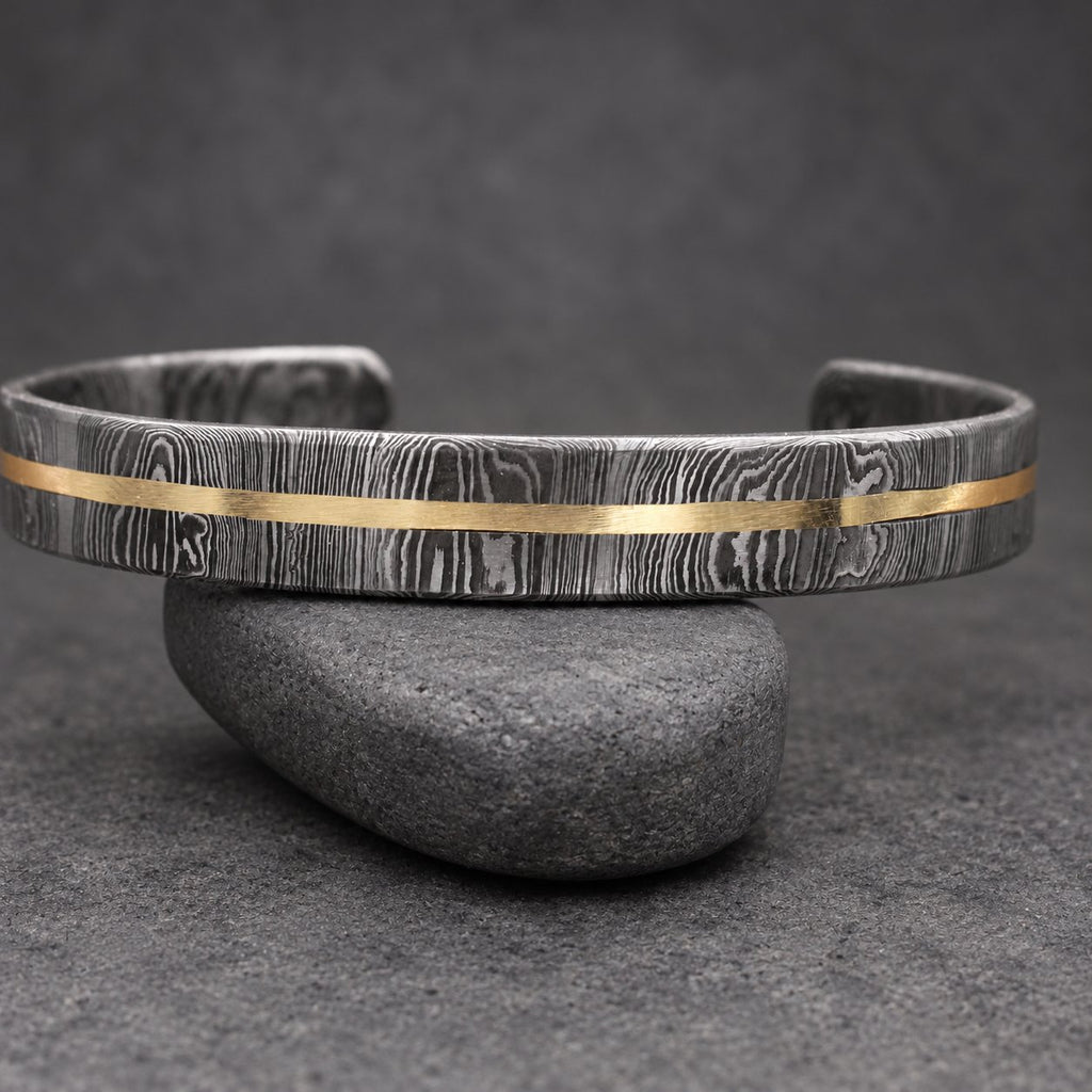 A Damascus steel cuff bracelet with a gold-colored stripe. The bracelet has a dark, textured pattern and is displayed on a wooden surface. This handcrafted jewelry piece is a stylish accessory.