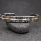 A Damascus steel cuff bracelet with a gold-colored stripe. The bracelet has a dark, textured pattern and is displayed on a wooden surface. This handcrafted jewelry piece is a stylish accessory.