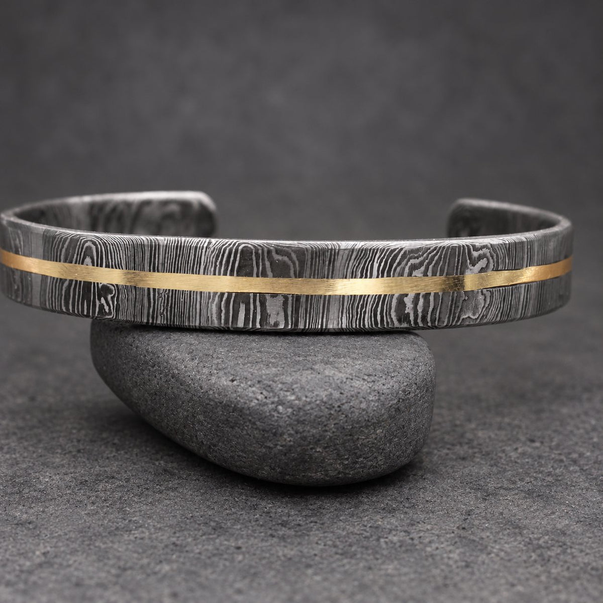 A Damascus steel cuff bracelet with a gold-colored stripe. The bracelet has a dark, textured pattern and is displayed on a wooden surface. This handcrafted jewelry piece is a stylish accessory.