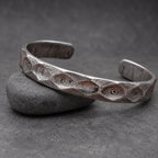 A silver cuff bracelet with a patterned surface. The open bracelet sits on a small, brown wooden object. The background is a wooden surface. The bracelet's design has a textured appearance.