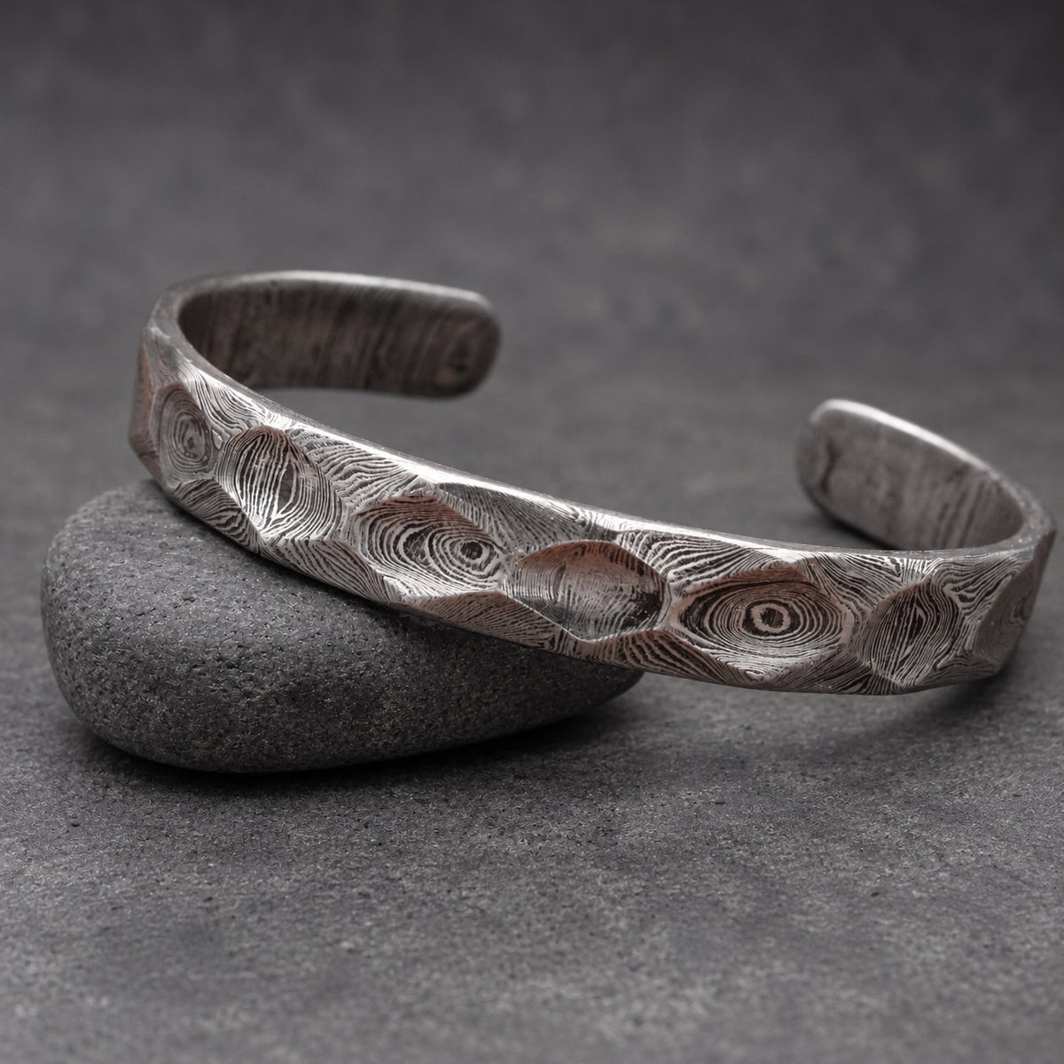 A silver cuff bracelet with a patterned surface. The open bracelet sits on a small, brown wooden object. The background is a wooden surface. The bracelet's design has a textured appearance.