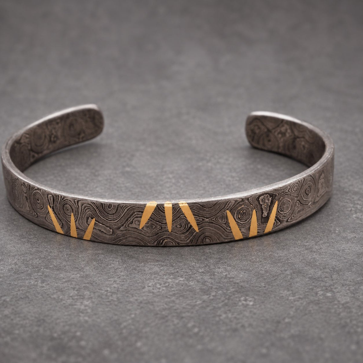 A silver cuff bracelet with a patterned texture and gold triangular details. The open-ended bracelet is displayed on a wooden surface. The jewelry item has a distinctive, artistic design, suitable for everyday wear.