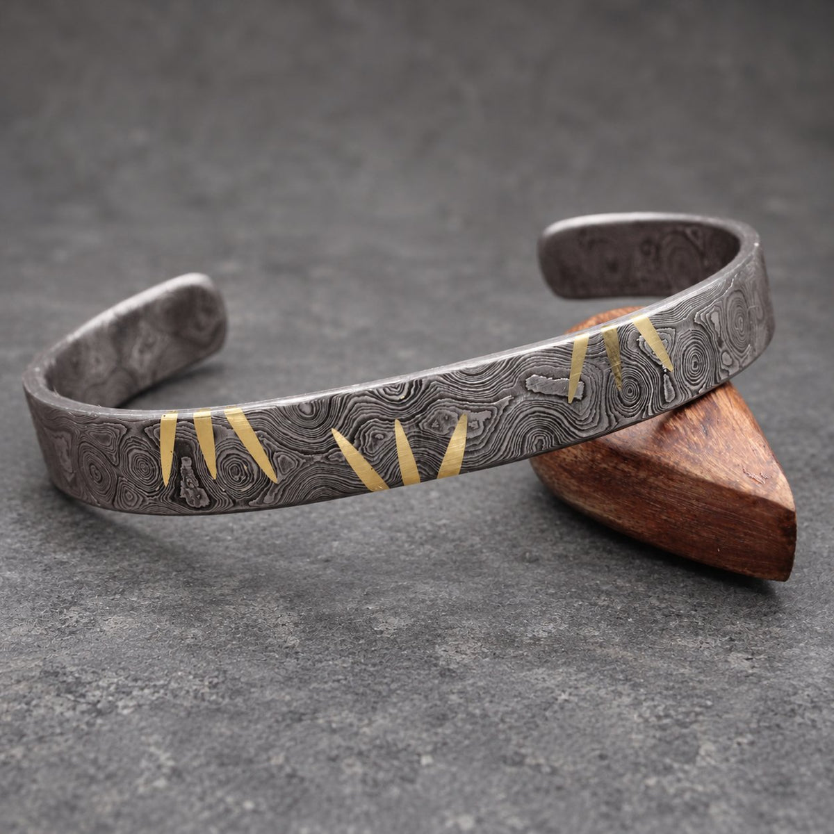 A silver cuff bracelet with a distinctive pattern and gold accents. The open-ended bracelet is displayed on a wooden stand against a wooden background. The gold accents are in a zig-zag pattern.