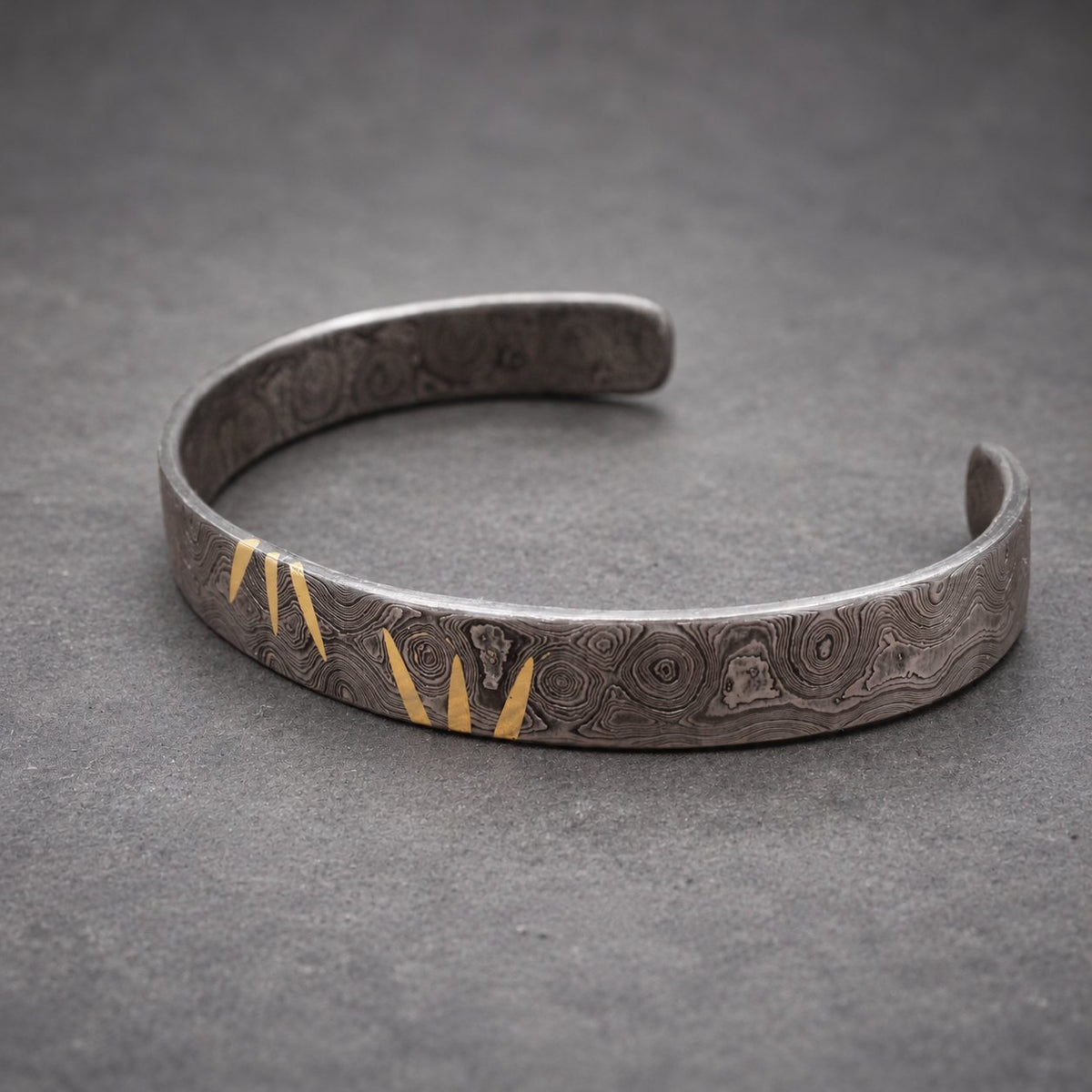 A silver-colored cuff bracelet with a unique pattern and gold-colored accents. The open-ended bracelet has a textured surface and a decorative design. The bracelet is displayed on a wooden surface.